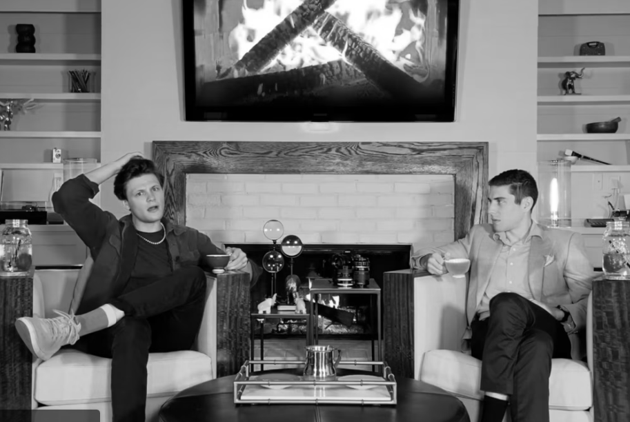 image of two men on chairs beside a fireplace, one is scratching his head in confusion